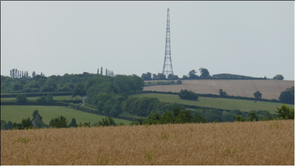 A tall mast, centre background with a wheat field in the foreground rolling down and then up again in a green valley