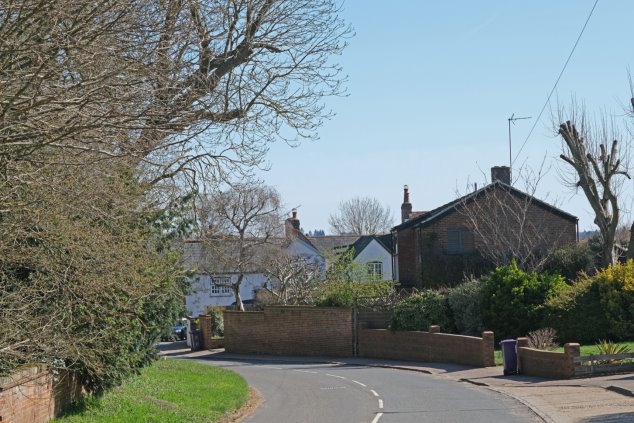 View 6 From the bend on Heath Lane looking east - bend in road with red brick wall and mature hedge on left and housing on right