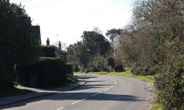 View 7 Entering Codicote village looking south-east along High Street - bend in the road with narrow grass verge and mature hedges on right and housing on left