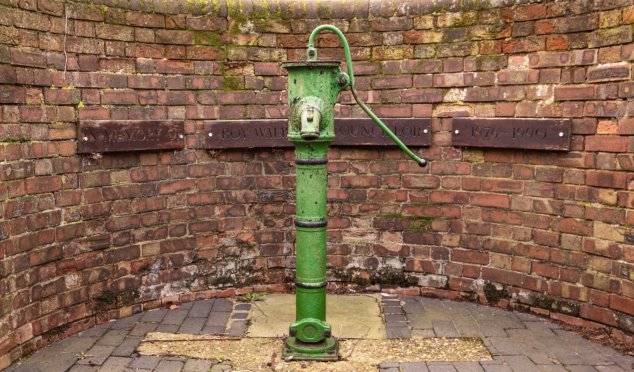 Old green painted iron water pump with handle set inside a semicircle red brick wall